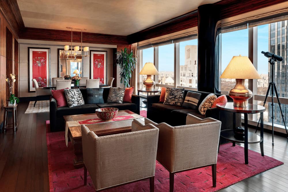 A large living room with multiple seating areas illuminated by stylish table lamps and contemporary chandelier. It has wide plank flooring and panoramic windows framing the city view.