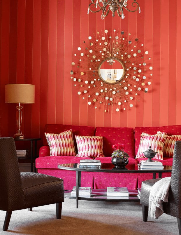 A sunburst mirror adds a gorgeous accent to the red striped wallpaper in this living room with skirted sectional and gray textured chairs surrounding the oval coffee table.