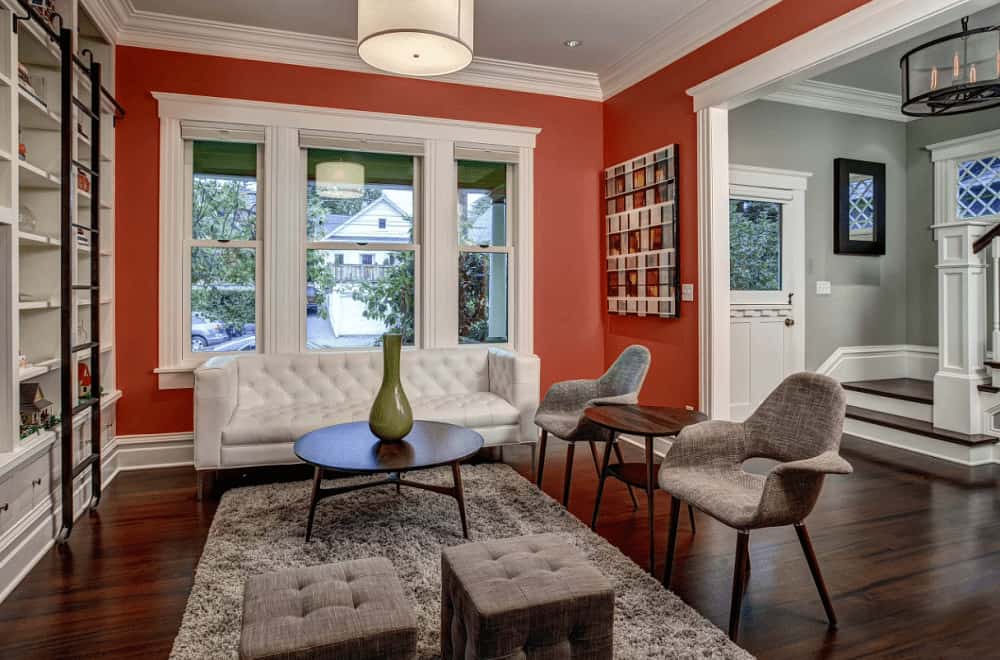 A red living room situated next to the gray foyer featuring cozy seats and a round coffee table topped with a green vase. It includes a drum pendant light and open shelving lined with a black ladder.