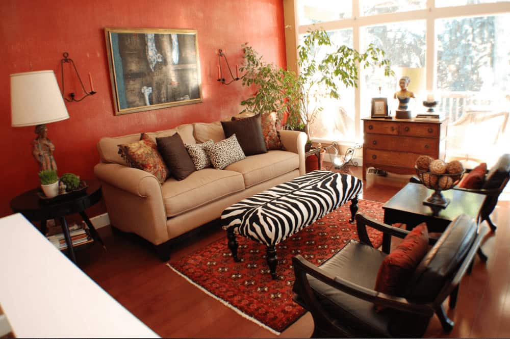 A zebra ottoman stands out in this red living room offering cozy seats and a wooden drawer that complements the smooth hardwood flooring topped by a red patterned rug.