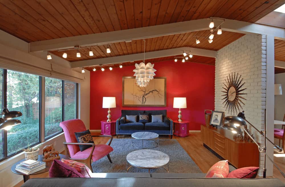 A pair of lovely floral pendant lights illuminate this living room along with track lights mounted on the wood beam ceiling. It is designed with a branch artwork and a sunburst clock fixed on the white brick wall.