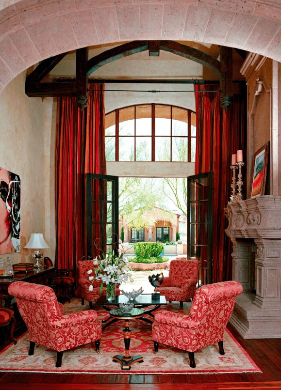 This living room boasts a concrete fireplace and black coffee table surrounded by red floral armchairs. It has wide plank flooring and a French door that opens to the yard.