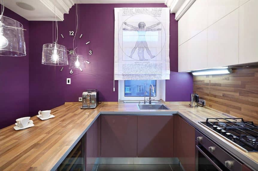 This small kitchen has a U-shaped peninsula to maximize the small space. This peninsula has a wooden countertop that matches with the backsplash of the cooking area. All of these elements are given a nice background of a purple wall adorned with a wall clock and decorative window shade.
