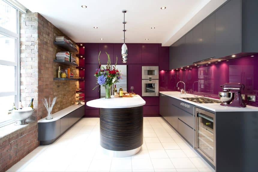 This kitchen is an eclectic fusion between modern and industrial-style. The red brick wall on one side has a window that brings in natural lights to the white ceiling and flooring tiles that is contrasted by modern dark gray cabinetry augmented by sleek dark purple walls and backsplash.