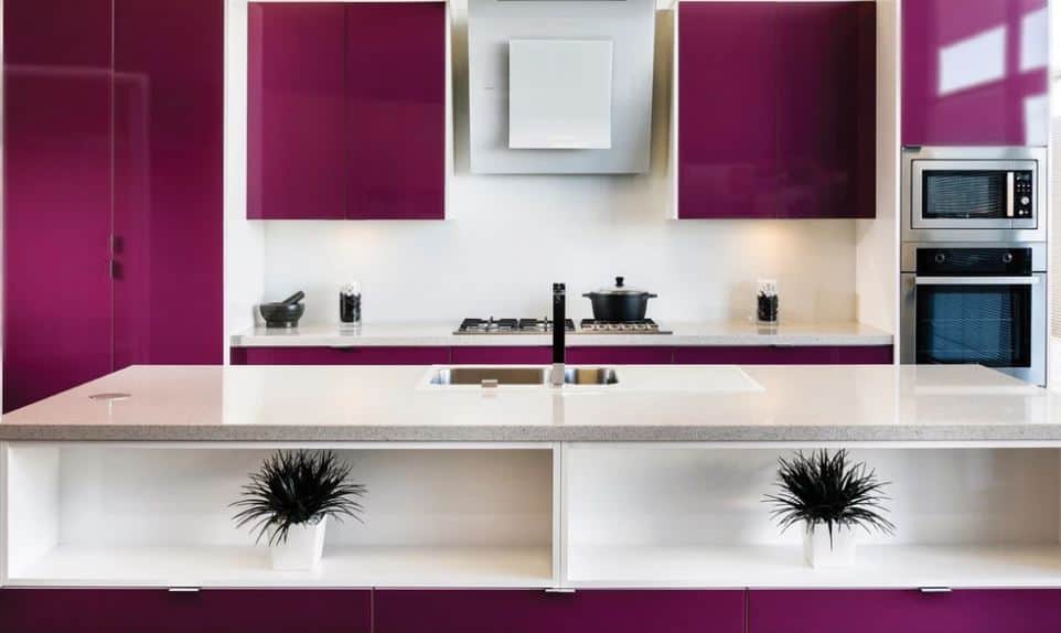 The cooking area of this kitchen has deep purple floating cabinets flanking the vent hood in the middle over the stove-top oven that has a white backsplash that contrasts the purple peninsula. Across from this is the sink area with the same countertop.