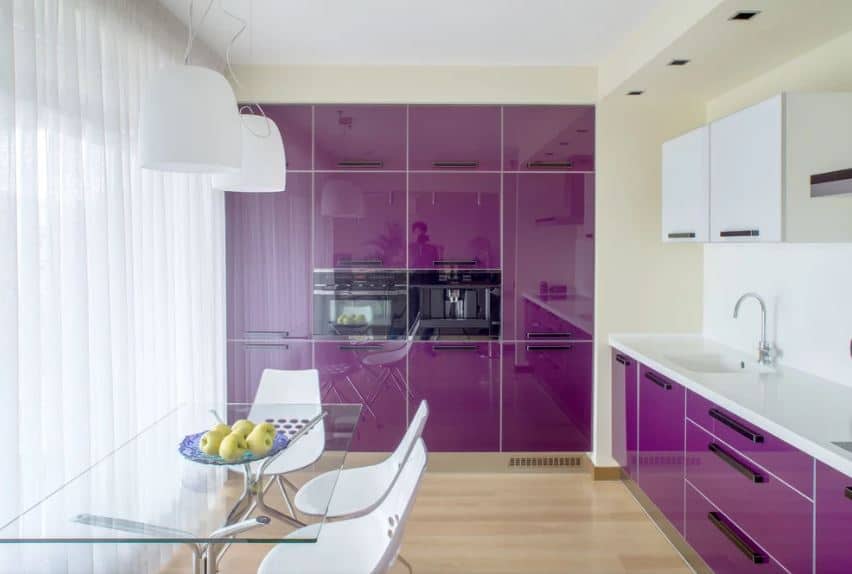The modern kitchen is also a part of the dining room that has a glass-top table and modern white chairs that are illuminated by the large curtained windows. These also brighten up the sleek deep purple cabinetry that are embedded into the walls of the kitchen.