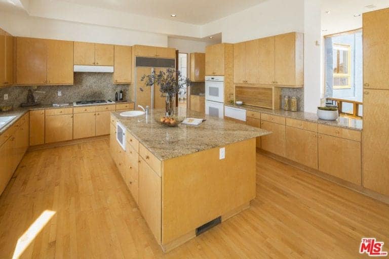 Brown kitchen offers white appliances and wooden cabinetry matching with the breakfast island that blends in with the hardwood flooring. It includes a built-in cooktop along with granite countertops and backsplash.