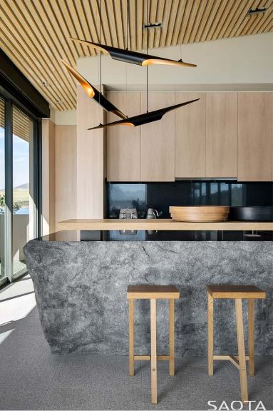 Wooden barstools sit at a boulder island with a raised wooden counter matching with the light wood cabinets that are contrasted by black granite backsplash. It is lighted by contemporary pendants that hung from the wood plank ceiling.