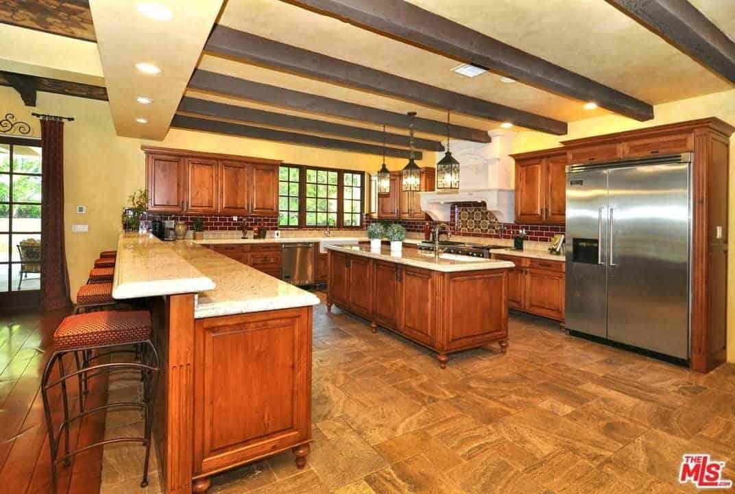 Lantern pendant lights hang over the granite top island that's fitted with an undermount sink and a gooseneck faucet. It is surrounded by wooden cabinetry and a two-tier peninsula lined with cushioned stools.