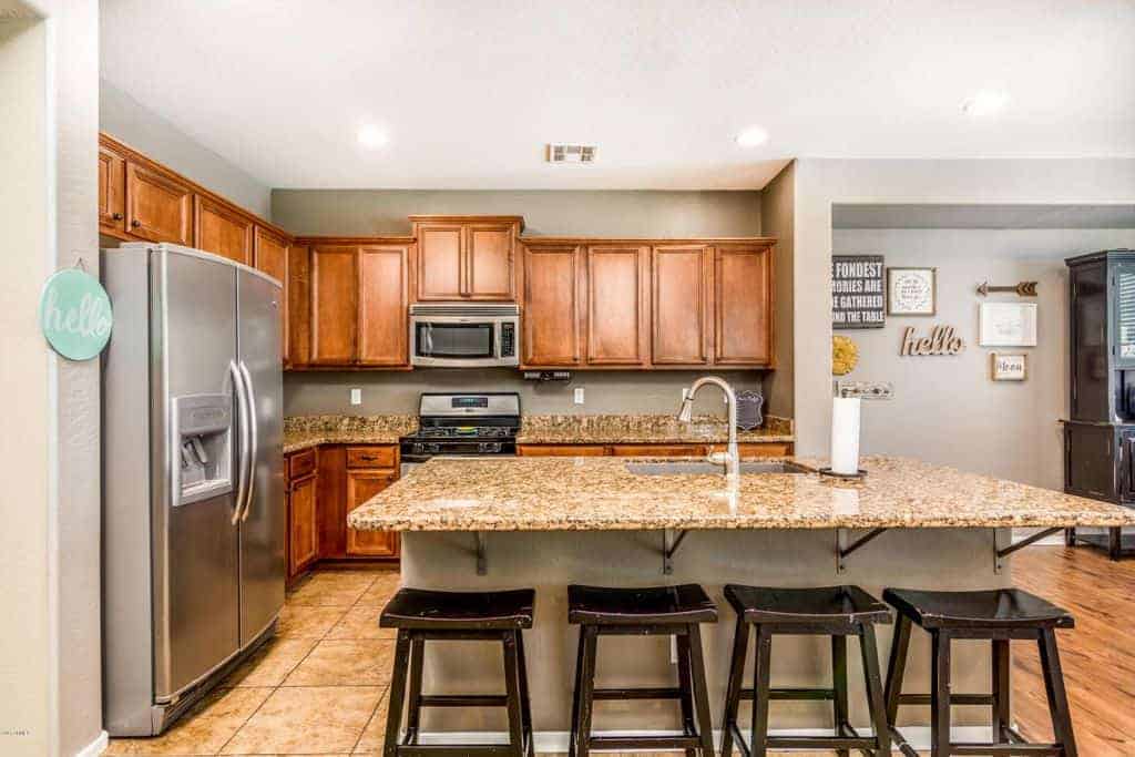 Black barstools sit at a taupe kitchen island that's topped with an undermount sink and a granite counter matching with the backsplash. It is accompanied by stainless steel appliances and wooden cabinetry.