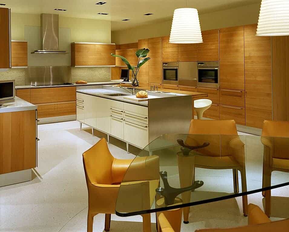 An eat-in kitchen with dome pendant lights and a glass top dining table surrounded by yellow leather chairs that complement the wooden cabinetry. It includes stainless steel appliances and two islands with quartz countertops.