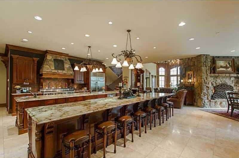 An open concept kitchen with dark wood cabinetry and two long island bars lighted by vintage chandeliers and recessed ceiling lights. It includes stainless steel appliances and a stone vent hood fixed against the black walls.
