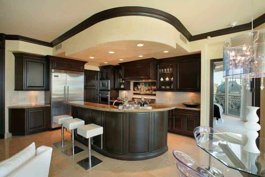Dark wood cabinetry matches the granite top breakfast island lined with modern white barstools. There's a glass dining set on the side lighted by a crystal drum chandelier.