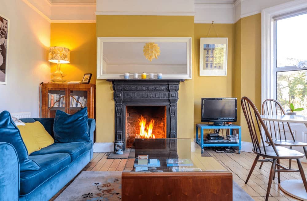 A blue velvet sofa stands out in this yellow living room with a glass top coffee table and white framed mirror mounted above the black fireplace.