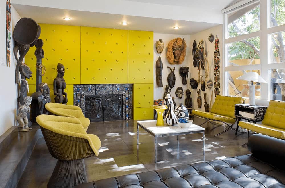 This living room is decorated with masks and sculptures sitting next to the gorgeous fireplace. It has yellow chairs and a black tufted sofa paired with a metal coffee table.