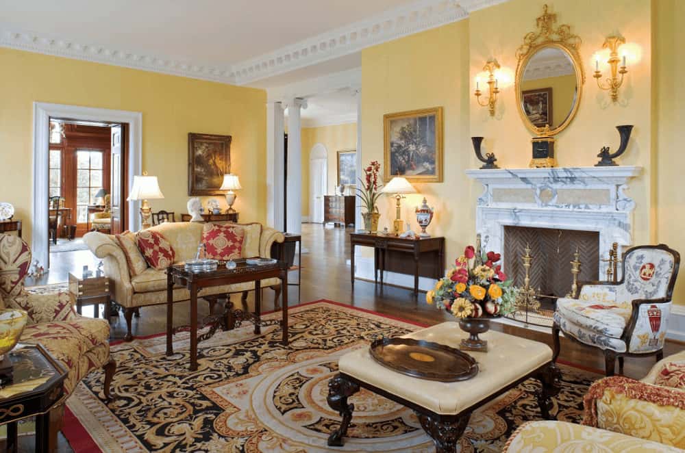 The elegant living room features classy seats and a chevron fireplace framed with white marble mantel. It includes a classic area rug and gorgeous ornate mirror mounted on the yellow wall lined with white crown molding.