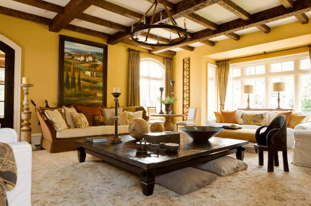 Mediterranean living room with a gorgeous landscape painting and a wrought iron chandelier that hung from the wooden coffered ceiling. It includes skirted seats and a large coffee table with gray pillows underneath.