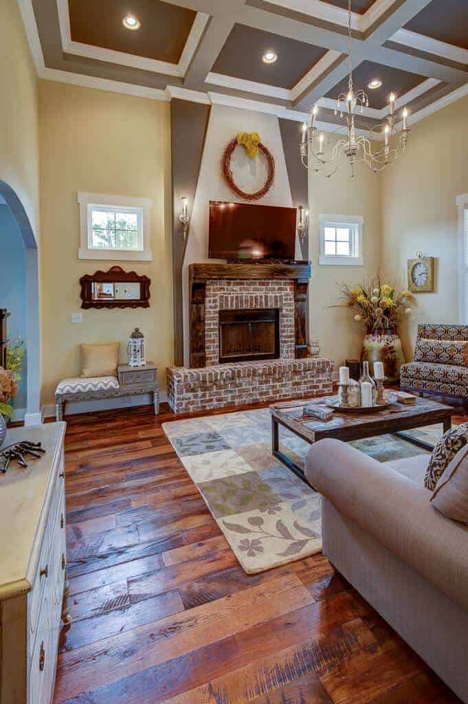 A candle chandelier illuminates this living room offering cozy seats and a brick fireplace framed with rustic mantel. It has a gray coffered ceiling and wide plank flooring topped by a floral checkered rug.