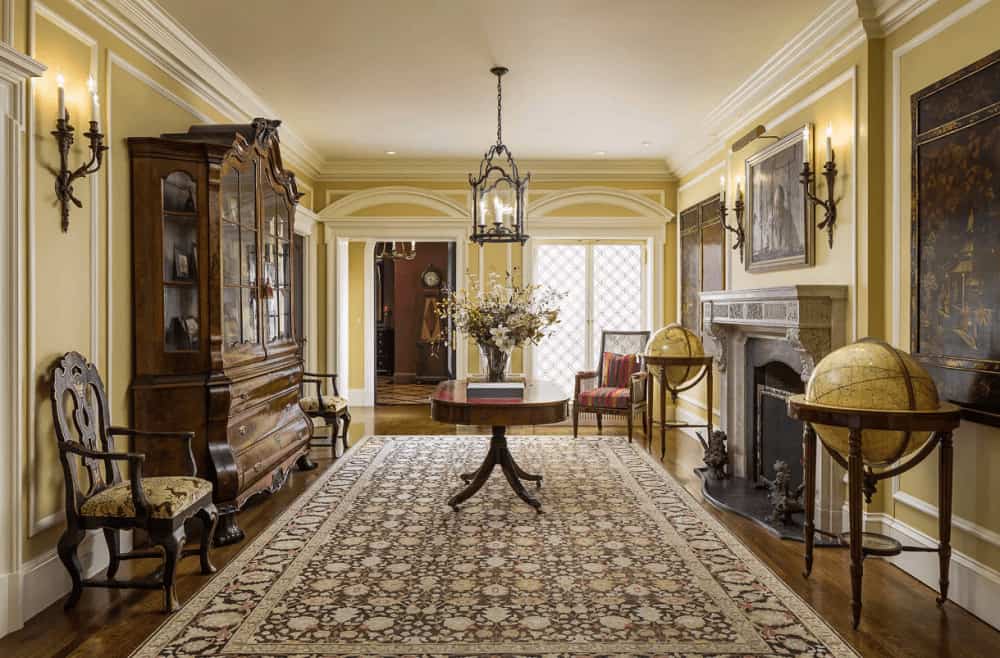The classic living room features a concrete fireplace and dark wood display cabinet flanked by cushioned armchairs. It is illuminated by a lantern pendant and candle sconces mounted on the yellow wainscoted walls.