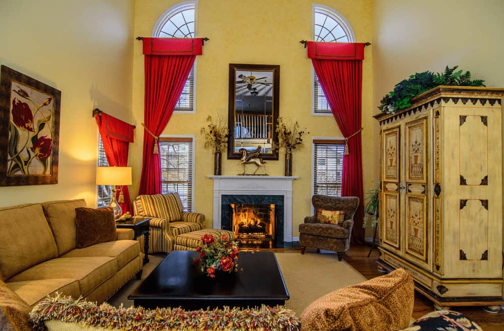 Classy red draperies covering the glazed windows stand out in this yellow living room with cozy seats and a fireplace topped with vases and horse sculpture.