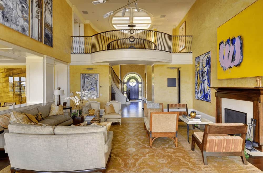 Yellow living room decorated with abstract paintings and a contemporary chandelier that hung from the high ceiling. It has plenty of seats and a glass enclosed fireplace framed with wooden mantel.