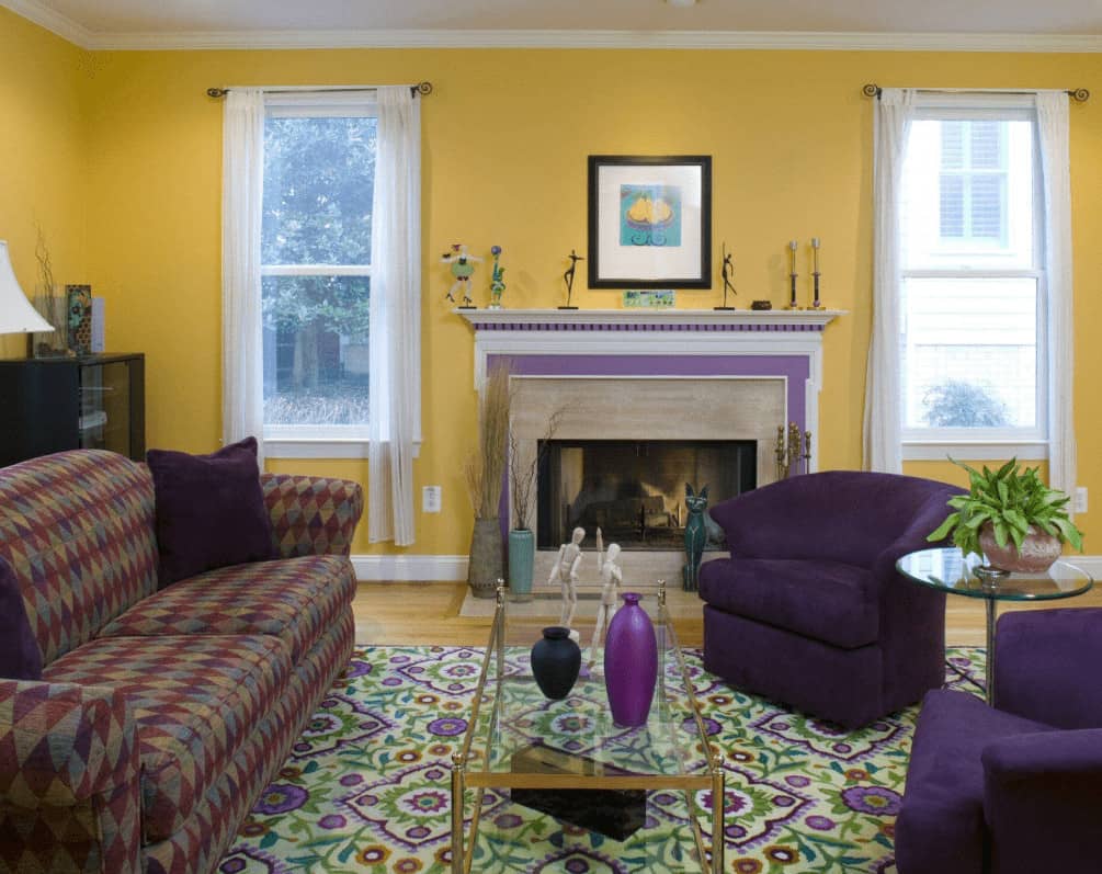 Yellow round back chairs add a nice contrast to the yellow walls fitted with white framed windows. This room has a patterned sofa and glass top coffee table along with a fireplace topped with decors and a black framed artwork.