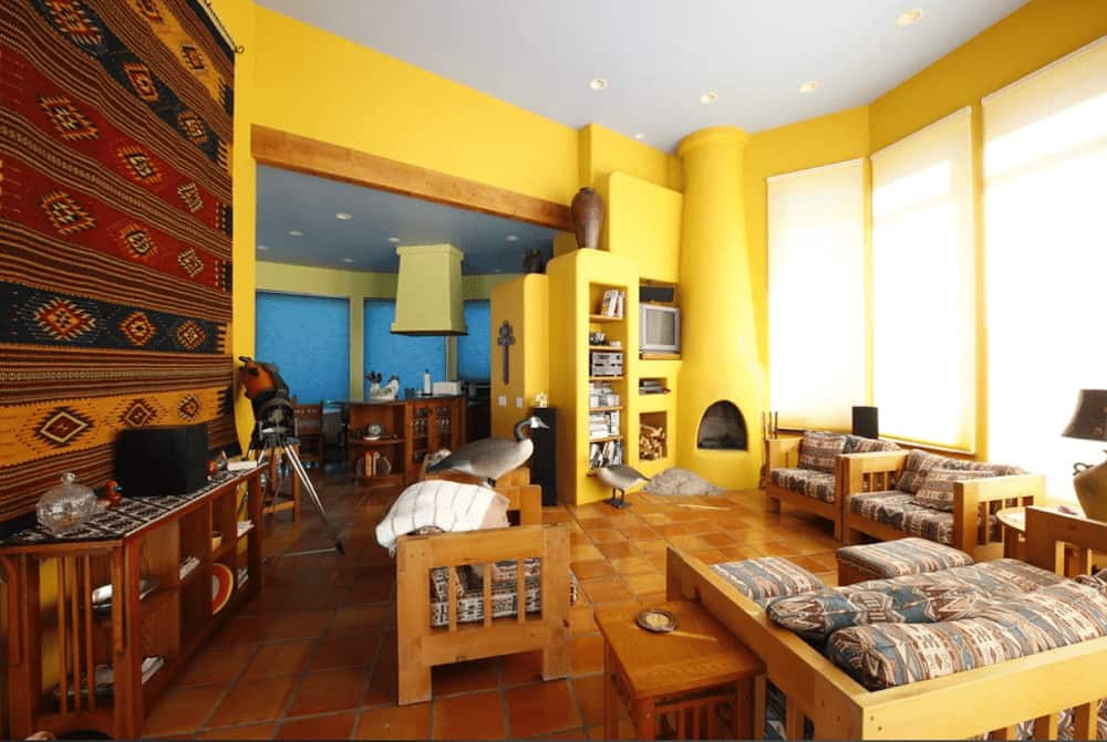 A patterned tapestry sets a gorgeous backdrop to the wooden console table in this yellow living room showcasing cushioned seats and a kiva fireplace with built-in shelving on the side.