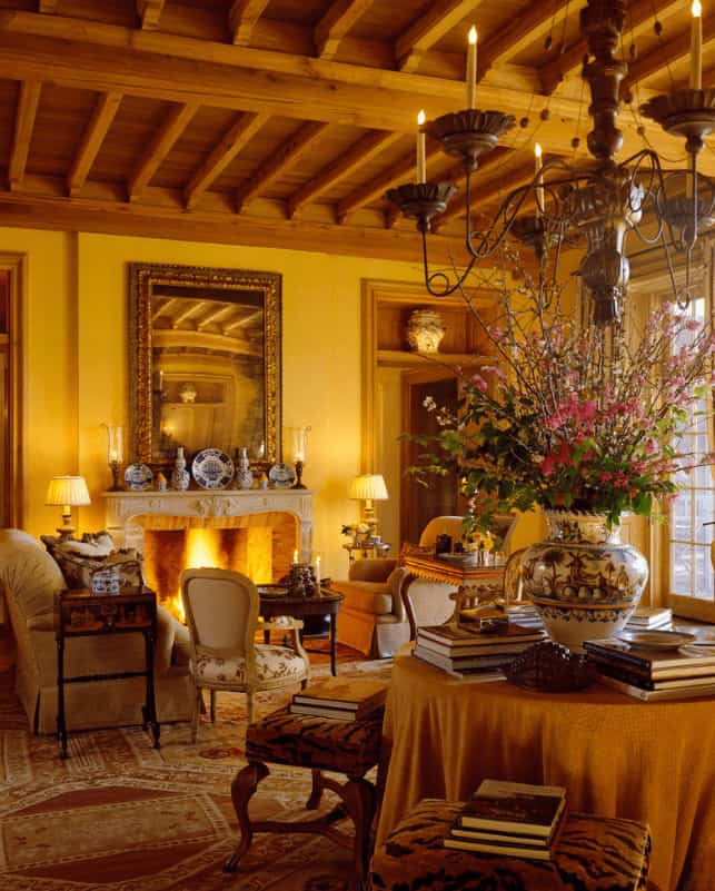 Warm living room with a fireplace and comfy seats over the printed area rug. It includes a skirted table surrounded with cushioned stools and lighted by a candle chandelier that hung from the wood beam ceiling.