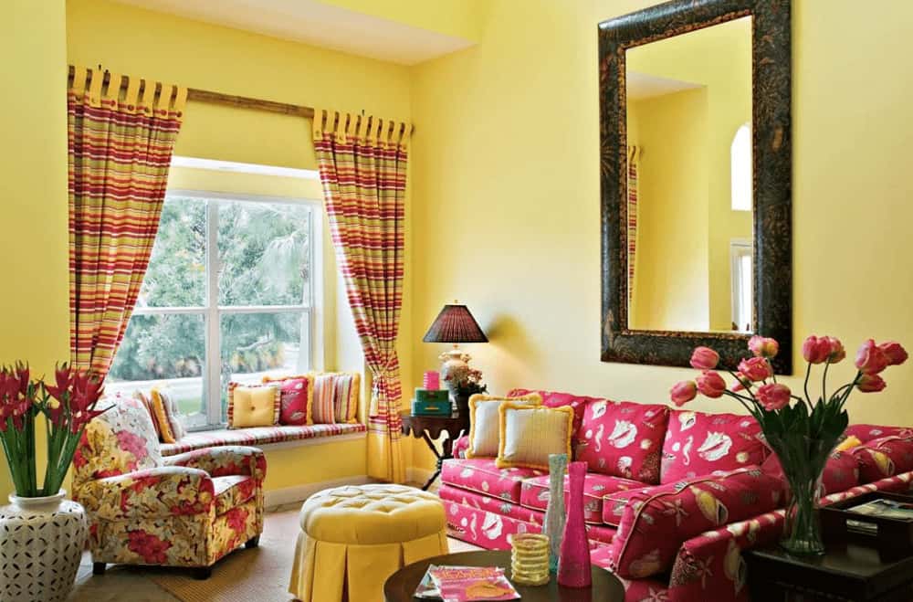 A large mirror hangs above the red sectional accompanied by a floral armchair and tufted round ottoman. There's a window seat nook on the side completed with small pillows and striped cushion that matches the draperies.