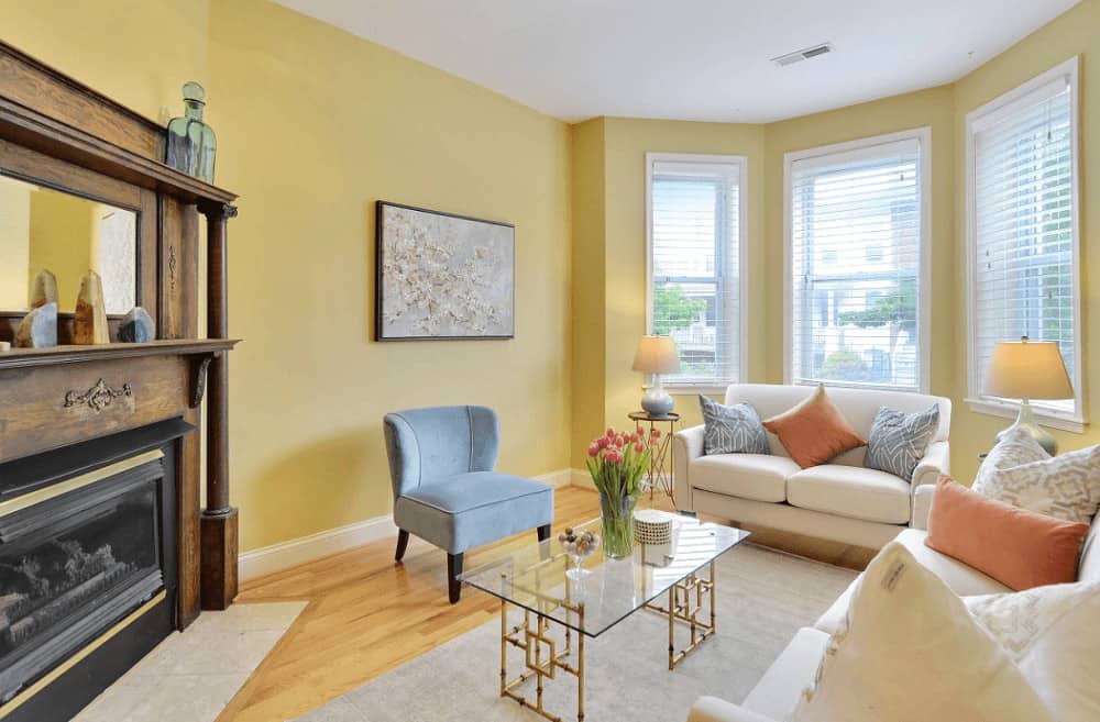 Bright living room with white sofas and a gray wingback chair paired with a stylish glass top coffee table. It includes lovely wall art and a corner fireplace with a mirror on top.