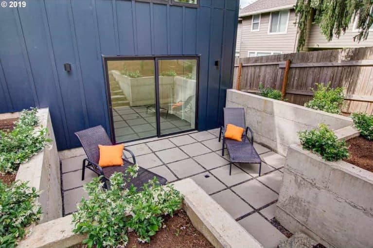 This is a small relaxation area at the back of the house accessed through glass sliding doors. It has gray concrete flooring and low walls that provide a view of the flowering shrubs planted by the edges that can be enjoyed on the couple of lounge chairs.