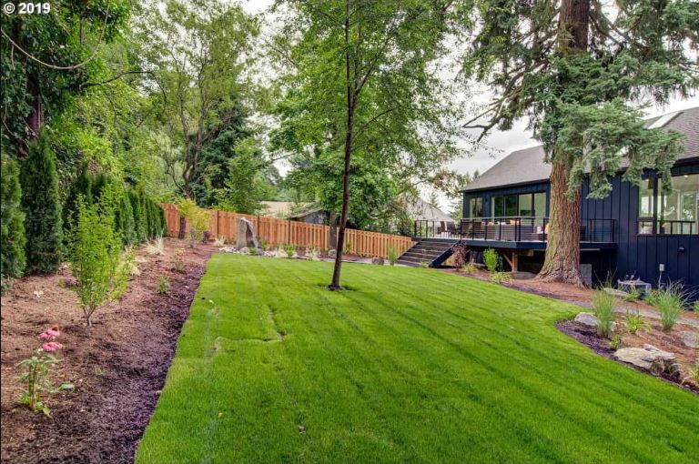 This simple green lawn at the backyard is surrounded by evenly spaced plants with shrubs and herbs in the mix. At the far end you can see the wooden fencing that surrounds the property and on the adjacent side to it is a row of tall shrubs that provide ample privacy.