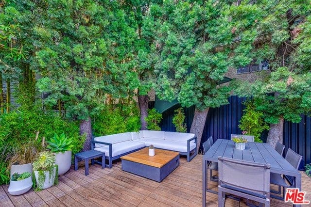 This backyard has a wooden plank flooring that complements the blue-gray wooden outdoor furniture of the outdoor dining area and the sitting area beside it that has an L-shaped wooden bench with white cushions that stand out against the background of green shrubs and tall trees.