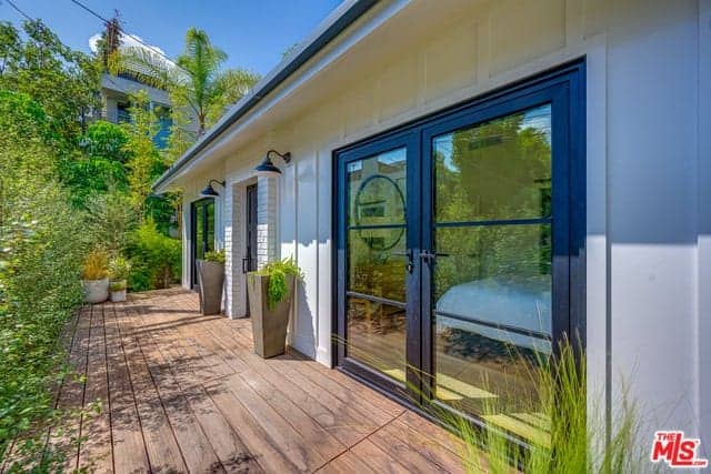The landscaping of this home has wooden plank walkways surrounding the house bordered with tall shrubs for privacy and has potted plants with tall pots that complement the white exterior walls that are lit with wall-mounted lamps.