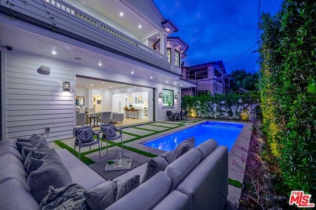 This backyard has a comfortable L-shaped gray sectional sofa at the sitting area beside the brilliant blue swimming pool. This wonderful area is surrounded by high white walls adorned with creeping plants and tall shrubs lit with yellow spotlights.