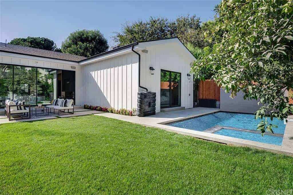 This is a backyard with a Farmhouse-style landscaping that has a simple beauty to its well-manicured lawn of grass and a blue pool that is surrounded by concrete walkways and wooden fences. There is also a sitting area with wooden armchairs that have white cushions.