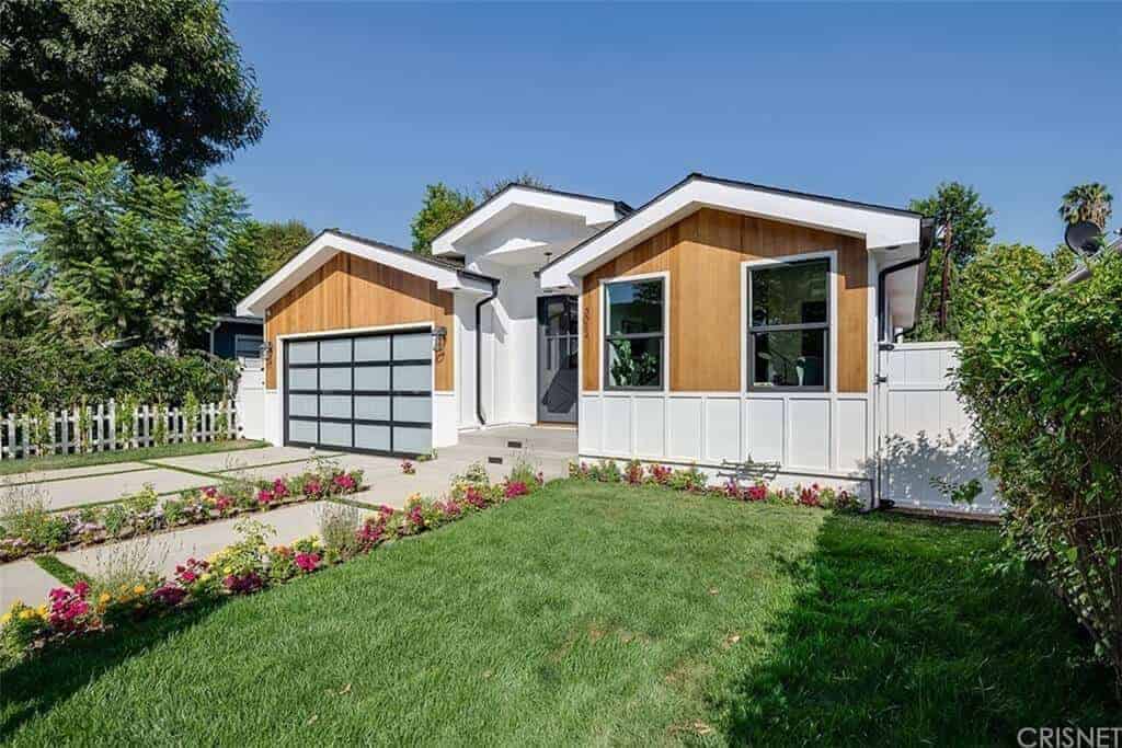 This is a charming view of the front yard of the house that has a Farmhouse-style landscaping. The front yard is divided into three sections. The left and middle side are the concrete driveway and walkway flanked with flowering shrubs and the right side is a lush lawn of grass.