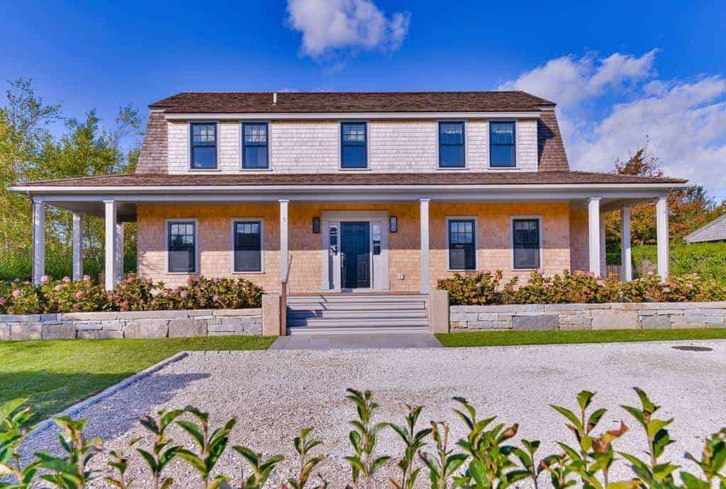 This is a Farmhouse-style landscaping that aims to complement and accent the home rather than compete for attention. There is a gravel driveway and walkway leading to a set of steps to a front porch that is adorned with a couple of ledges on both sides bearing flowering shrubs that complements the pink exterior walls.