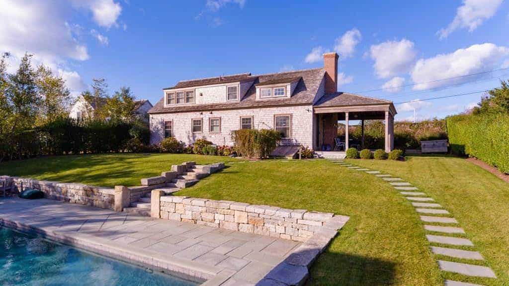 This lovely wooden Farmhouse-style home is perfectly framed by this landscape scenery. There is a nice blue sky above that complements the well-manicured lawn of grass that has a couple of couple of concrete walkways. One walkway leads to a pool that is surrounded with stone and concrete while the other on goes around.