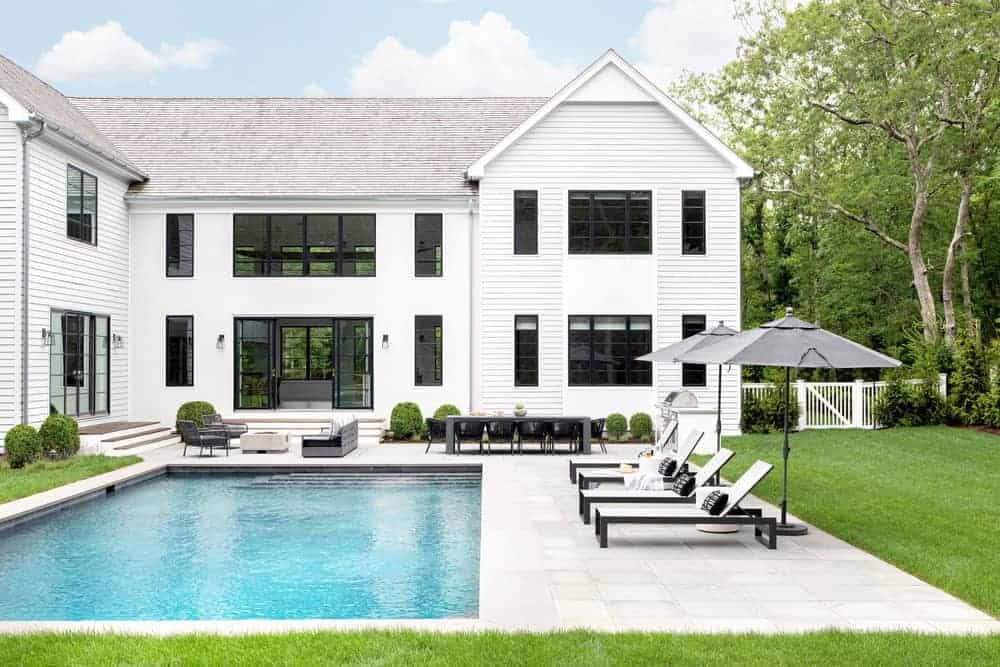 This backyard rectangular pool is paired with white lawn chairs shaded by the beach umbrellas on the concrete floor surrounding the pool. These flooring also houses a sitting area and a dining area near the white exterior walls.