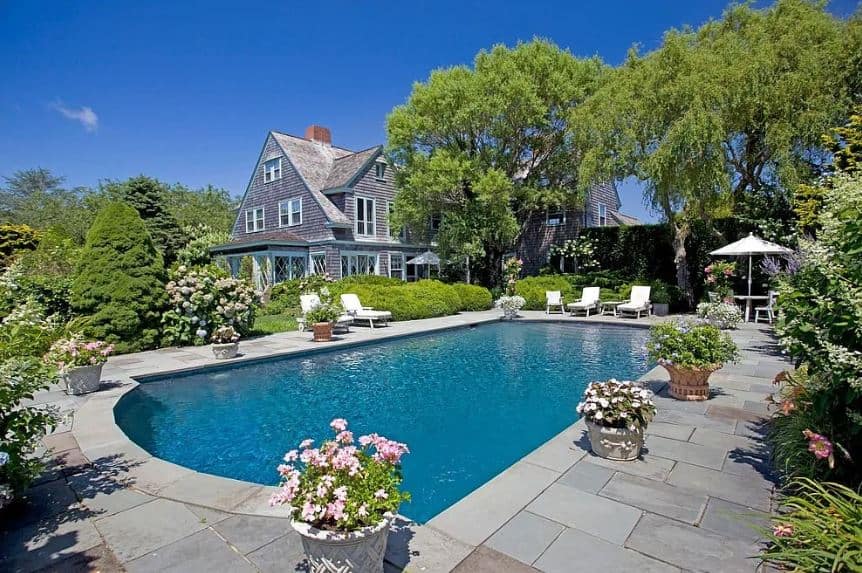 The charming pool is surrounded by potted flowering plants that pairs well with the surrounding flowering shrubs that encroach on the concrete walkways around the pool that has several sitting areas and lounge chairs to enjoy the sun.