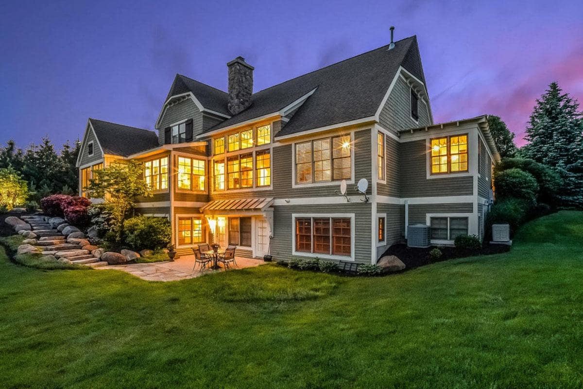 This is a backyard view of the house. You can see here that it is filled with warm yellow lights cascading from its wide glass windows that offer a view of the lush green landscaping surrounding the house with a lovely stone walkway on the side leading to a sitting area lit with warm yellow lights.