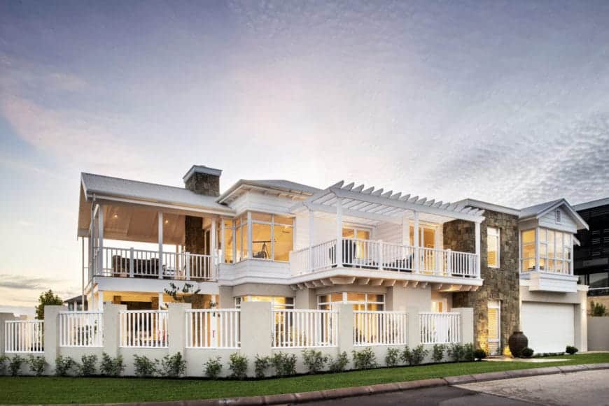 This large home is dominated by its light hues of white railings and windows paired with the beige walls almost unnoticeable with the yellow lights. This is mirrored by the fence surrounding the property. It has beige columns and white railings complemented by the carpet of grass just outside the fence.