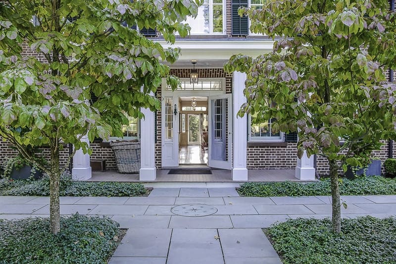 The exterior walls of this house is dominated by the red brick walls that is contrasted by the white pillars and complemented by the surrounding green landscaping. There is a gray concrete walkway that is flanked by shrubs and medium sized trees.