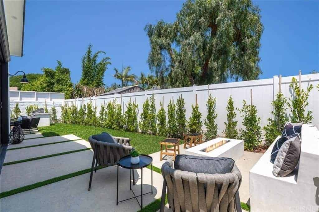 This backyard has a warm and comfortable sitting area due to its modern firepit that is inlaid with white concrete matching the stone bench. This is paired with a couple of gray outdoor chairs that stand out against the concrete flooring that leads to a small lawn of grass and end with a surrounding white concrete wall adorned with shrubs.