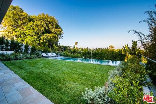 This is an amazing piece of backyard with a Farmhouse-style landscaping. The property is surrounded by tall shrubs that provide fencing and privacy for those enjoying the lawn of well-maintained grass and the pool on the far side with white lawn chairs.