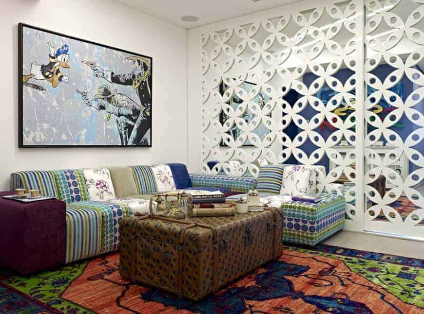 This living room boasts a stylish mirrored wall and an L-shaped sofa with large wall art on top paired with a chest coffee table over a brightly colored rug.