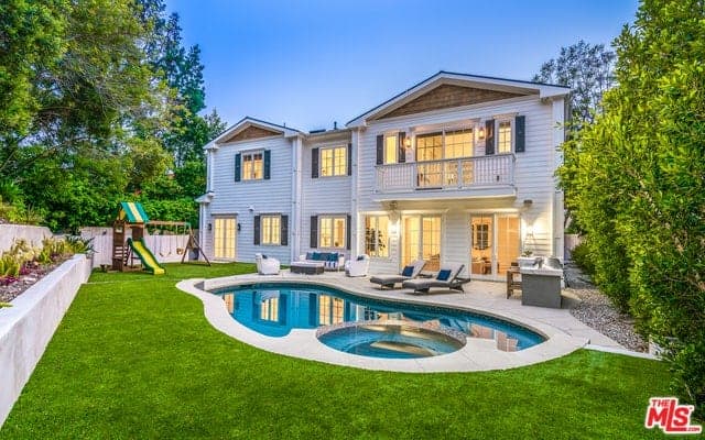 This view of the house shows a sitting area with white furniture, a couple of lawn chairs and an outdoor cooking area. These areas look down on the unique pool surrounded by a lush green lawn and a small playground for the kids on the side.