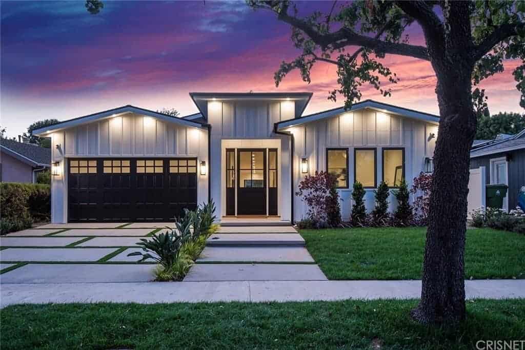 The gray concrete walkway and driveway both have green accents that emphasize the lines of the concrete blocks. These leads to the house lit with warm yellow lights of the wall-mounted lamps that works well with the well-manicured lawn of grass and its shrubberies.