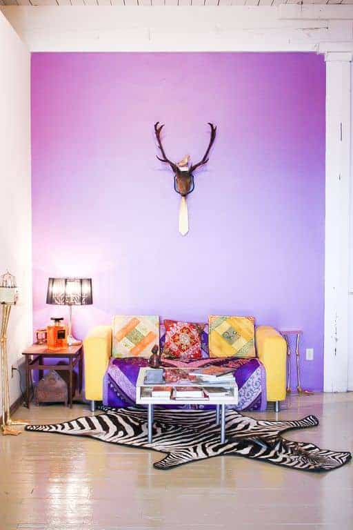 Eclectic living room with sleek coffee table and a yellow couch that adds a striking contrast to the purple accent wall mounted with a stag head decor. It includes black lampshade and zebra rug that lays on the hardwood flooring.